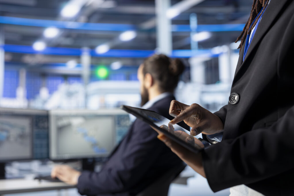Professional using a digital tablet while monitoring operations in a modern facility, with colleague working at computer in the background.